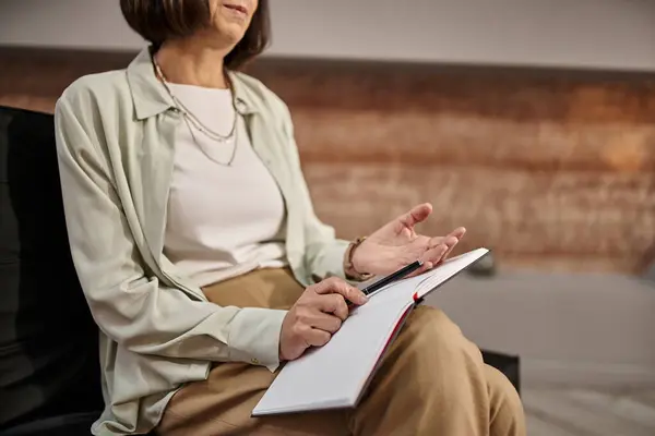 Cropped view of middle aged psychologist sitting with notebook and pen in consulting room — Stock Photo