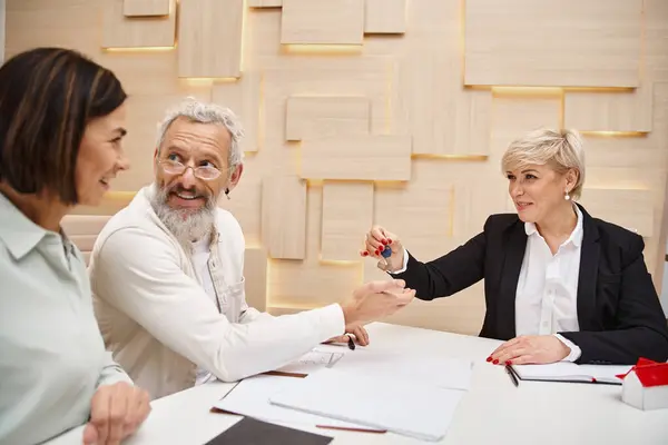 Agente inmobiliario de mediana edad dando la llave de la casa nueva a la pareja casada feliz en la oficina de bienes raíces - foto de stock