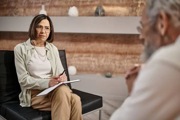 Psicólogo de mediana edad en gafas sentado y hablando con el hombre barbudo durante la sesión de terapia - foto de stock