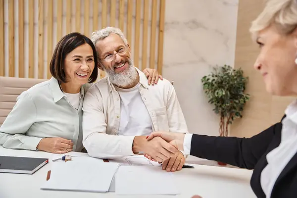 Щасливий чоловік середнього віку тремтить руками з ріелтором біля дружини і робить угоду в офісі з нерухомості — стокове фото