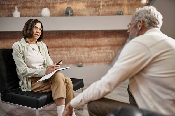 Psicologo di mezza età in occhiali seduto e parlando con l'uomo barbuto durante la sessione di terapia — Foto stock