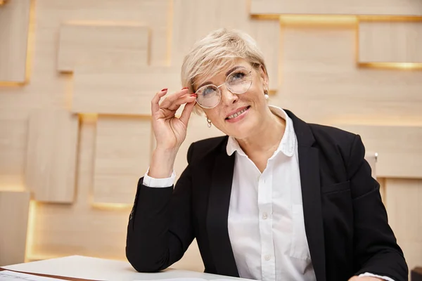 Щаслива блондинка середнього віку ріелтор в окулярах працює в офісі нерухомості, дивлячись на камеру — стокове фото