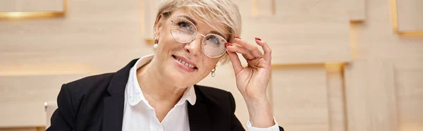 Щаслива блондинка середнього віку ріелтор в окулярах дивиться на камеру в офісі нерухомості, банер — стокове фото