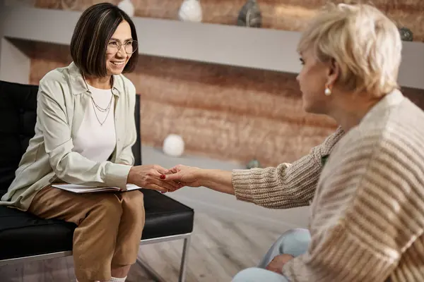 Psychologue d'âge moyen dans des lunettes souriant et serrant la main avec une femme blonde après la session — Photo de stock