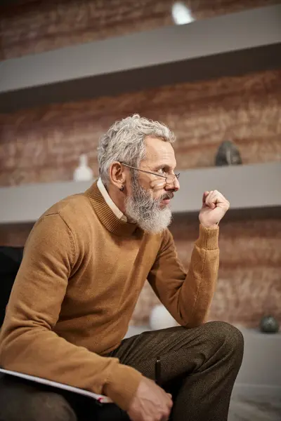 Bearded middle aged psychologist in glasses holding pen near notebook and listening during session — Stock Photo