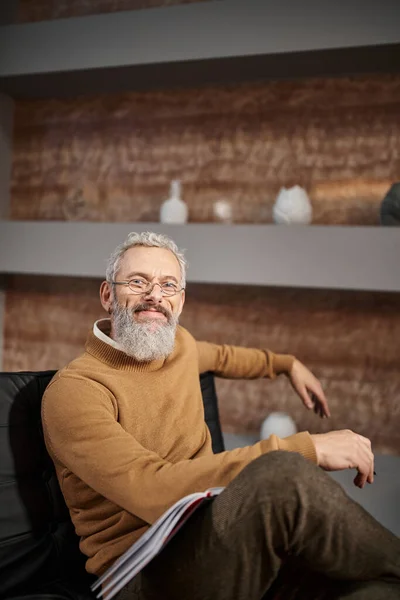 Cheerful middle aged psychologist in glasses smiling and sitting on armchair during therapy session — Stock Photo