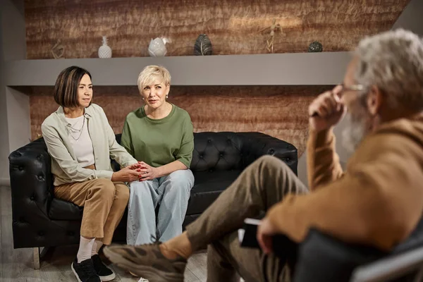 Middle aged lesbian couple talking and sitting on couch near psychologist during session — Stock Photo