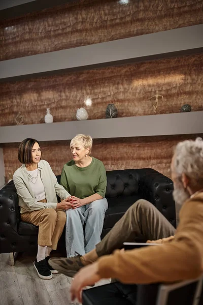 Pareja lesbiana casada de mediana edad hablando y sentada en el sofá cerca del psicólogo durante la sesión - foto de stock