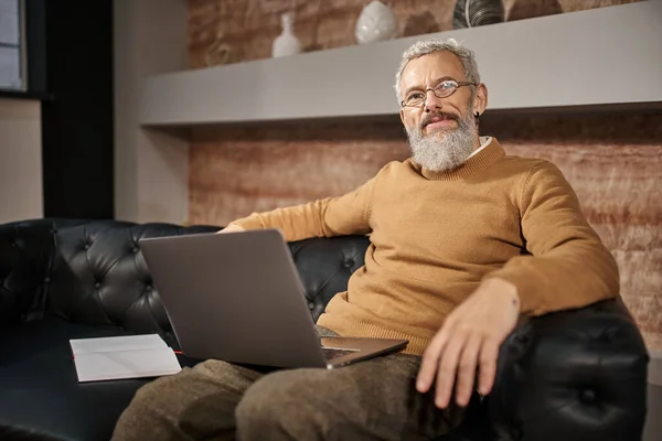 Psicólogo alegre de mediana edad con barba sentado en el sofá de cuero con el ordenador portátil, consulta en línea - foto de stock