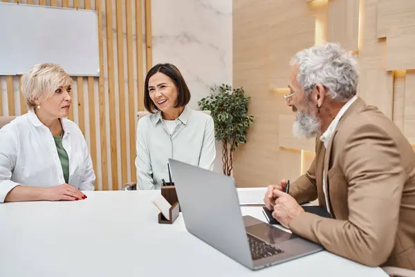 Happy middle aged multicultural lgbt couple sitting next to bearded realtor in real estate office — Stock Photo