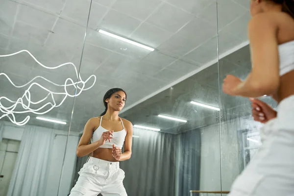 Affascinante ballerina afroamericana in top sportivo bianco e pantaloni che regolano i capelli lunghi in studio moderno — Foto stock
