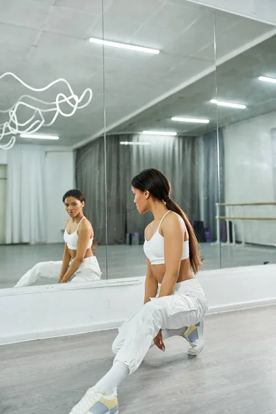 Artistic african american dancer in white sportswear stretching near mirror in studio, choreography — Stock Photo