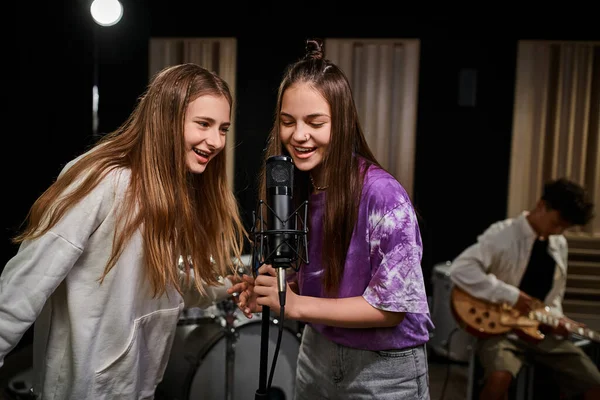 Allegre ragazze adolescenti che cantano felicemente mentre il loro amico suona la chitarra sullo sfondo, gruppo musicale — Foto stock