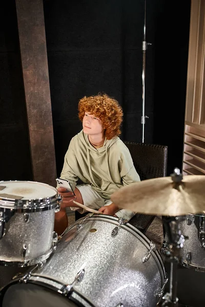 Allegro ragazzo dai capelli rossi adolescente in abbigliamento comodo che tiene il telefono cellulare prima di suonare la batteria in studio — Foto stock