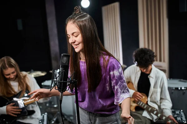 Cheerful brunette teenage girl singing happily with her friends playing instruments in studio — Stock Photo