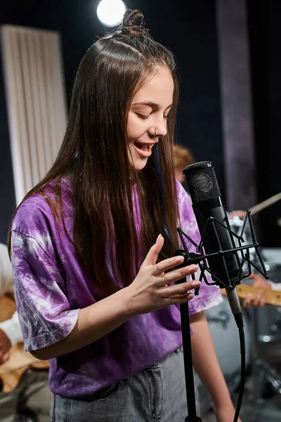 Vertical shot of adorable teenage girl singing next to her blurred guitarists, musical group — Stock Photo