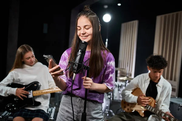 Focus on cheerful teenage girl singing and looking at her phone next to her blurred guitarists — Stock Photo