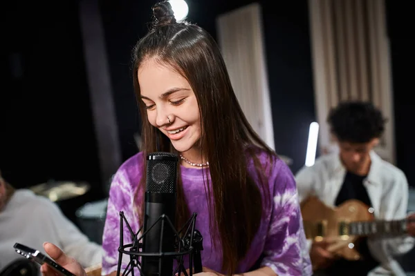 Jolly cute teenage girl with piercing singing and looking at mobile phone next to her friends — Stock Photo