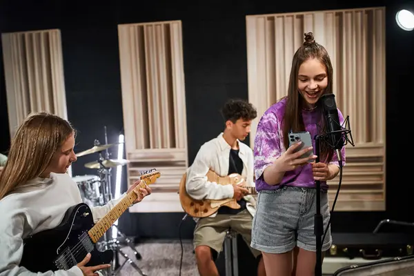 Cheerful teenage girl looking at phone and singing next to her friends playing guitars, musical band — Stock Photo