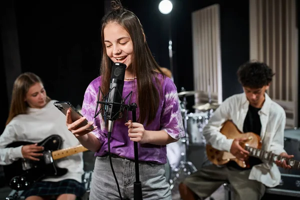 Focus on jolly teenage girl in vivid attire singing and looking at phone near her blurred guitarists — Stock Photo