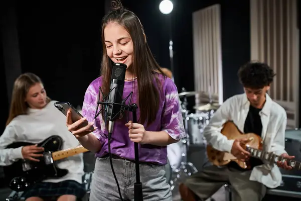 Se centran en la alegre adolescente en traje vívido cantando y mirando el teléfono cerca de sus guitarristas borrosos - foto de stock