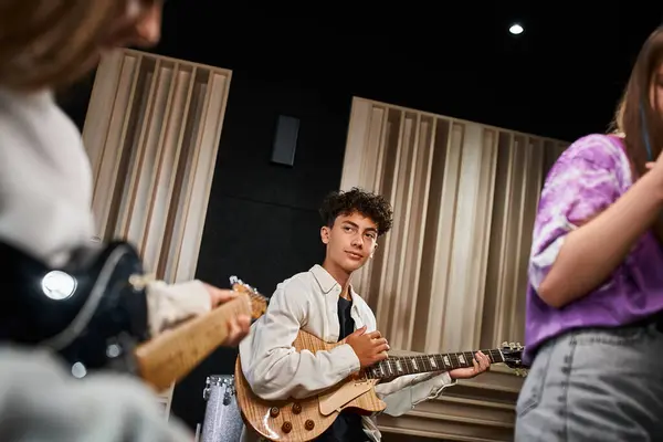 Concentrado lindo adolescente sosteniendo la guitarra y mirando a su borrosa banda de miembros en el estudio - foto de stock