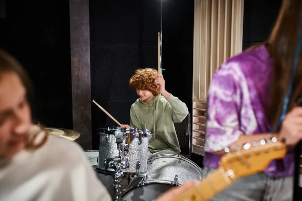 Concentre-se no menino adolescente de cabelos vermelhos em traje casual tocando sua bateria perto de seus membros da banda turva — Fotografia de Stock