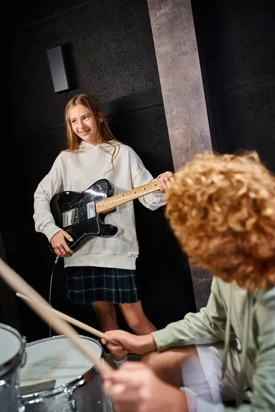 Foco na menina adolescente alegre em traje casual tocando guitarra ao lado de h er desfocado baterista de cabelos vermelhos — Fotografia de Stock