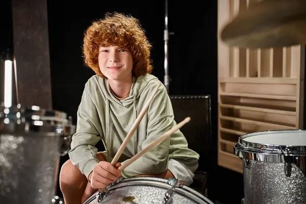 Cheerful adorable red haired teenager in casual vivid attire in front of drum set looking at camera — Stock Photo
