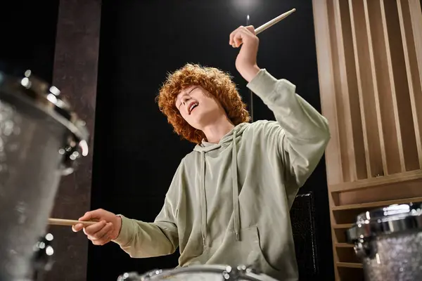 Bonito alegre adolescente menino com cabelo vermelho em traje casual tocando sua bateria ativamente enquanto no estúdio — Fotografia de Stock