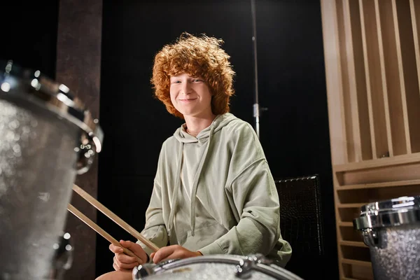 Cheerful adorable teenager with red hair in casual outfit in front of his drum set smiling at camera — Stock Photo