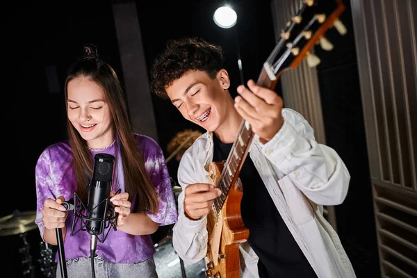 Bien parecido adolescente alegre con aparatos ortopédicos tocando la guitarra junto a su lindo vocalista en el estudio - foto de stock