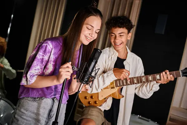Adorável alegre adolescente cantando música ao lado de seu amigo com aparelho tocando guitarra em estúdio — Fotografia de Stock