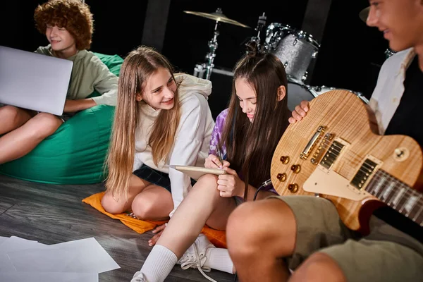 Alegres meninas adolescentes bonitos em roupas vívidas escrever letras perto de seus amigos com laptop e guitarra — Fotografia de Stock