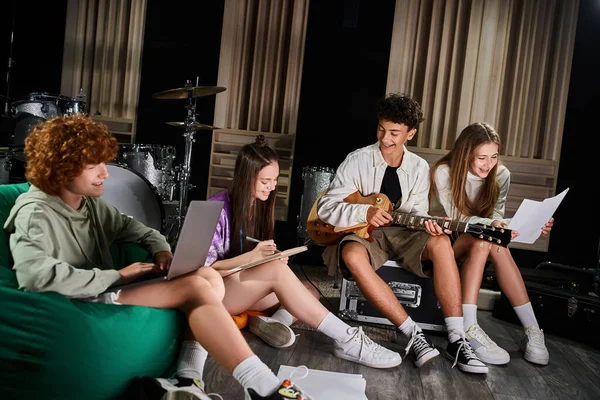 Vier fröhliche Teenager-Bandmitglieder in lässiger Kleidung sitzen mit Gitarre und Laptop und schreiben Texte — Stockfoto