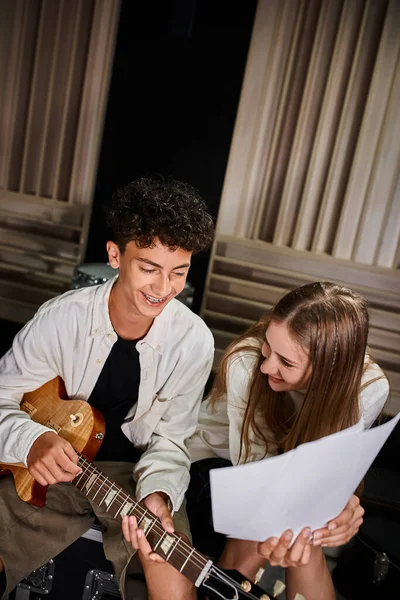 Joyous adorable teenage friends in casual attires playing guitar and looking at lyrics, musical band — Stock Photo
