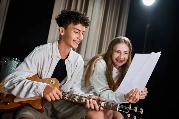 Cheerful teenage boy with braces and blonde girl playing guitar and looking at lyrics in studio — Stock Photo