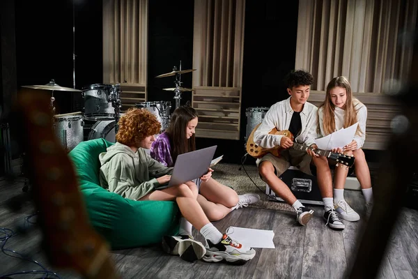 Fröhliche niedliche Teenager-Freunde sitzen mit Laptop und Gitarre auf dem Boden und schauen sich im Studio die Texte an — Stockfoto