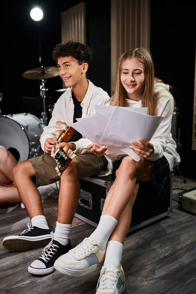 Alegre adolescente olhando para letras ao lado de seu amigo com aparelho tocando guitarra, grupo musical — Fotografia de Stock