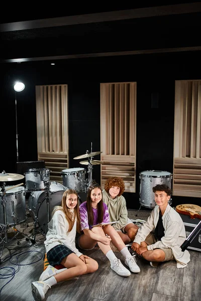 Talentosos adolescentes alegres sentados en el estudio con letras y mirando a la cámara, grupo musical - foto de stock
