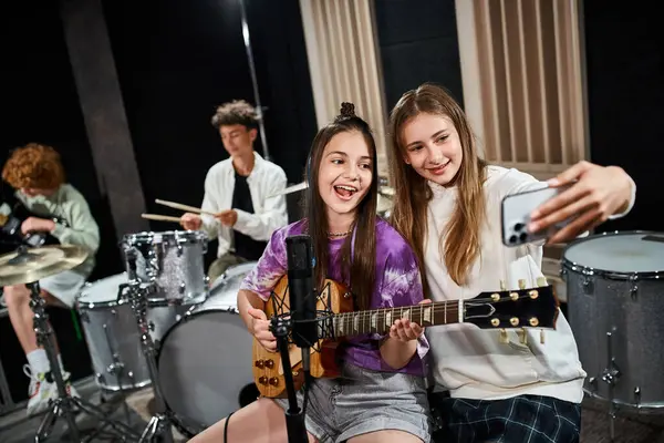Focus on jolly teenage girls taking selfie and singing with blurred friends playing instruments — Stock Photo