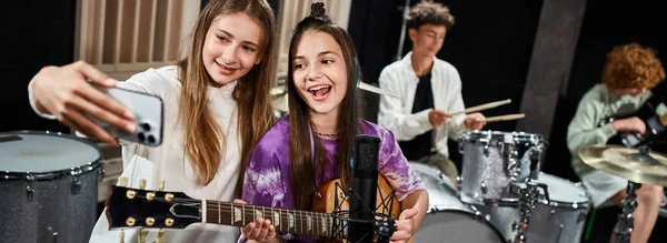 Focus on happy teens taking selfie with blurred friends playing instruments on backdrop, banner — Stock Photo
