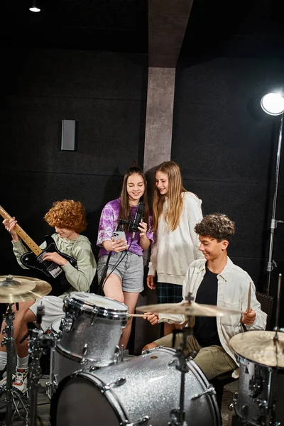 Fröhliche Teenager-Mädchen singen, während Jungen im Studio Schlagzeug und Gitarre spielen, Musikgruppe — Stockfoto