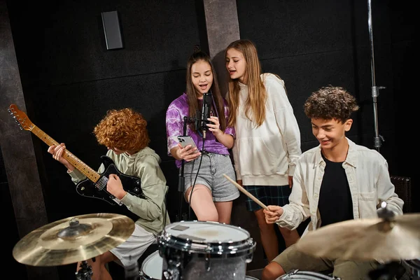 Chicos adolescentes lindos en atuendos casuales tocando la batería y la guitarra, mientras que las niñas cantan canción, grupo musical - foto de stock
