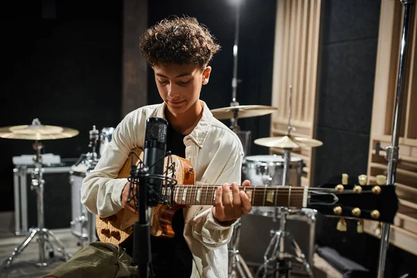 Веселий талановитий хлопчик-підліток у стильному повсякденному вбранні грає на гітарі в студії і злегка посміхається — стокове фото