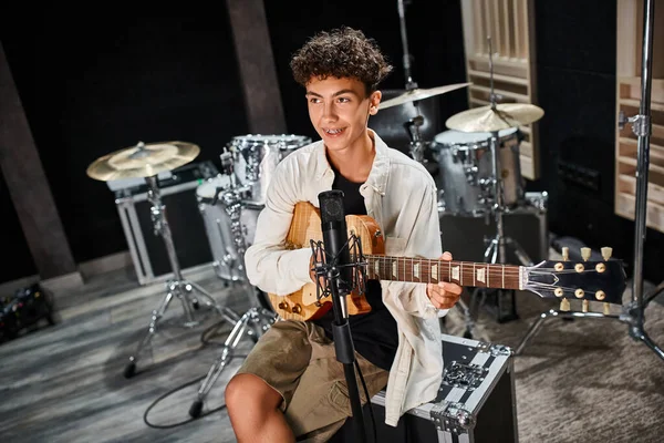 Радісний чарівний хлопчик-підліток у повсякденному вбранні з брекетами грає на гітарі і дивиться вбік — стокове фото