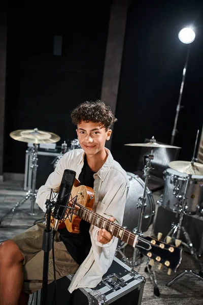 Cheerful talented teenage boy in casual outfit playing his guitar and looking at camera in studio — Stock Photo