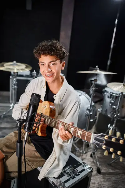 Buen aspecto músico adolescente con aparatos ortopédicos en traje casual tocando la guitarra y sonriendo a la cámara - foto de stock