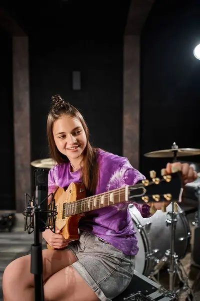 Alegre guitarrista adolescente en traje casual vívido tocando la guitarra frente al micrófono en el estudio - foto de stock