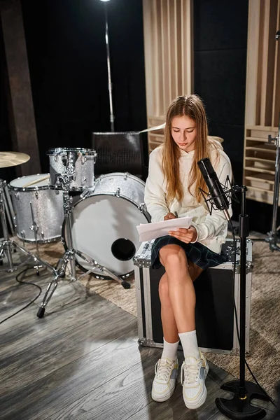 Pretty blonde teenage girl in casual attire sitting in front of microphone and looking at lyrics — Stock Photo